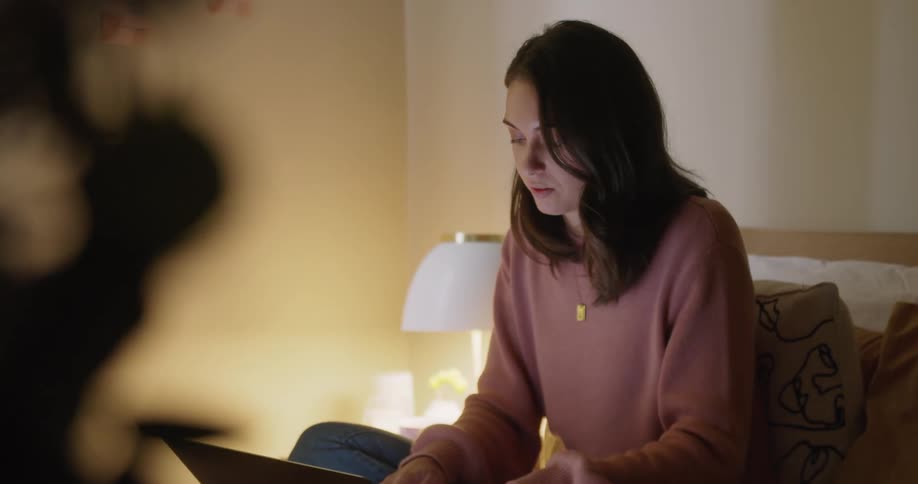 A woman sits on a bed working on her laptop