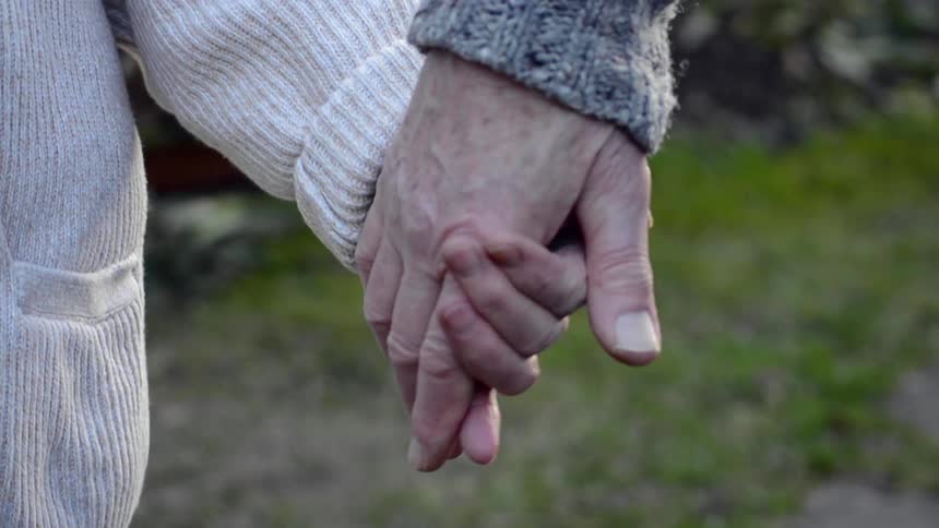 An elderly man and woman reach their hands out to one another and hold hands