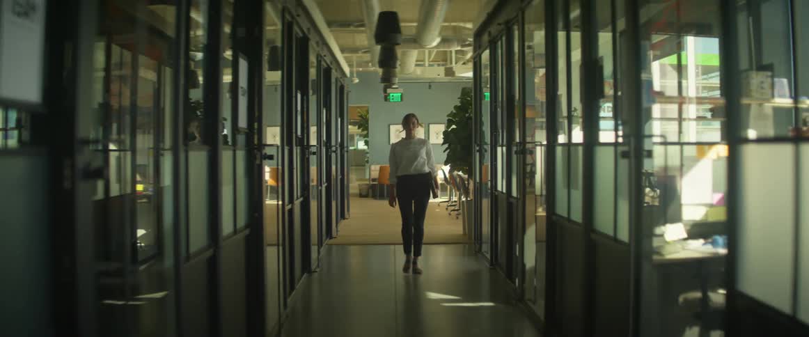 A woman in a white blouse confidently walks down an office hallway