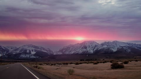 The sun sets over a snow capped mountain range near a desert as lightning strikes