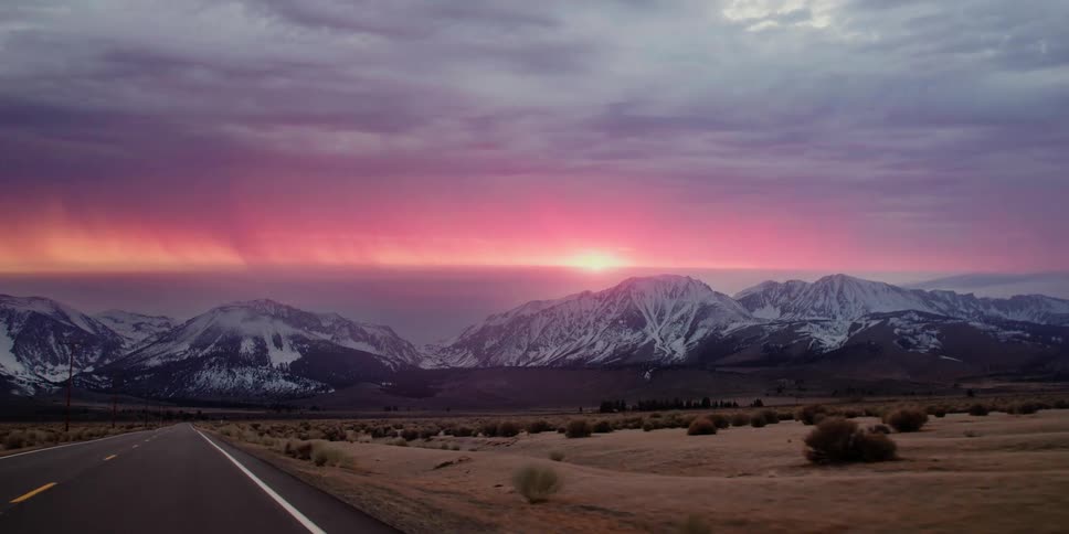 The sun sets over a snow capped mountain range near a desert as lightning strikes