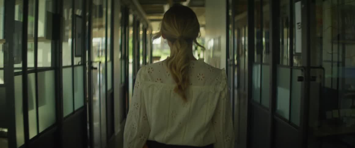 Blonde woman walks along corridor past glass walled offices into empty work area