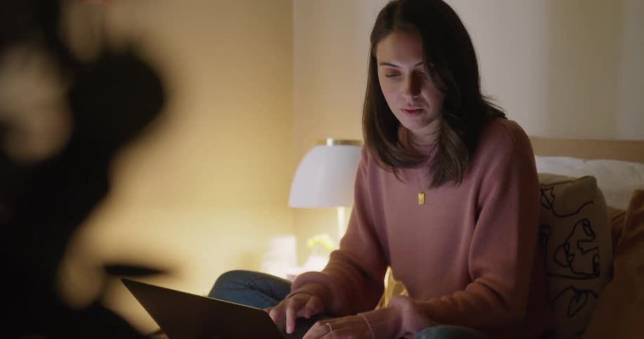 A woman sits on her bed and types on her laptop