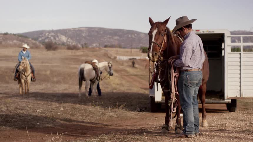 A man in a cowboy hat fastening the reins on a horse in a countryside