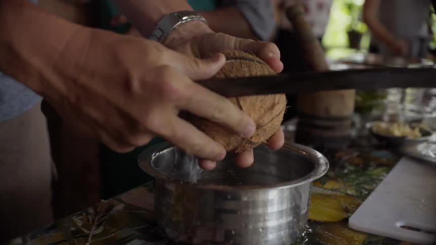 A white male cuts open a coconut with a large machete