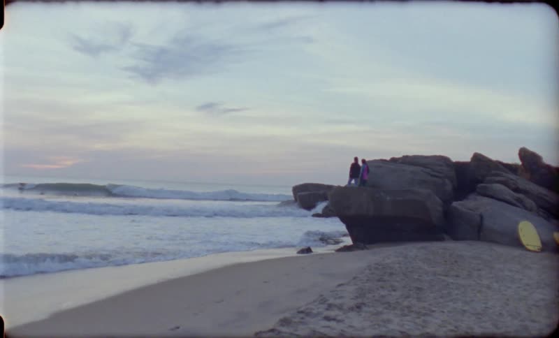 Woman and man stand on a big rock and watch the foamy waves at dusk