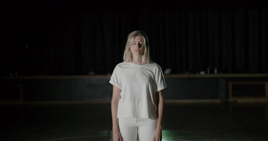 A white woman dressed all in white stands in a room with her eyes closed