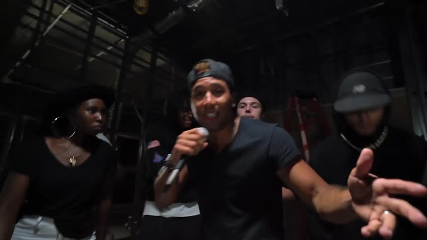 A man holding a microphone raps and gestures as a group of people behind him dance to the beat