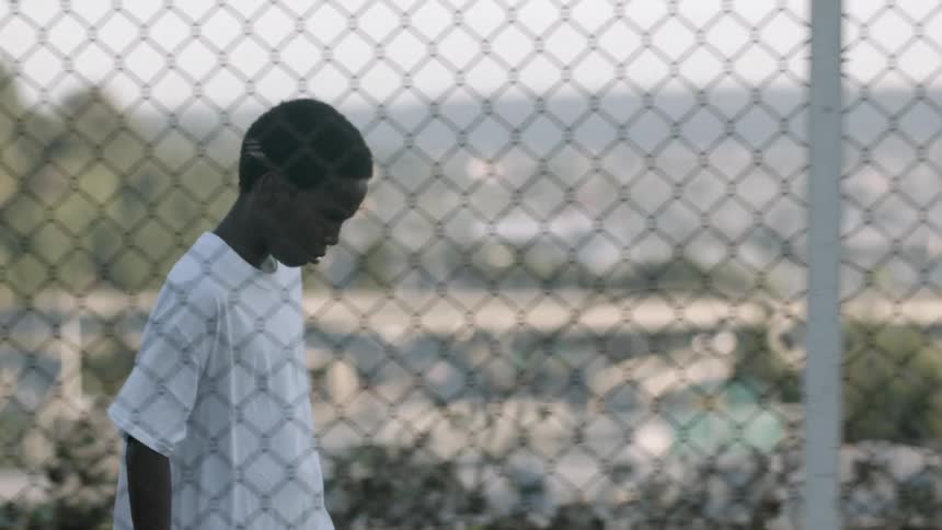 Young boy walks behind wire fence near city at day