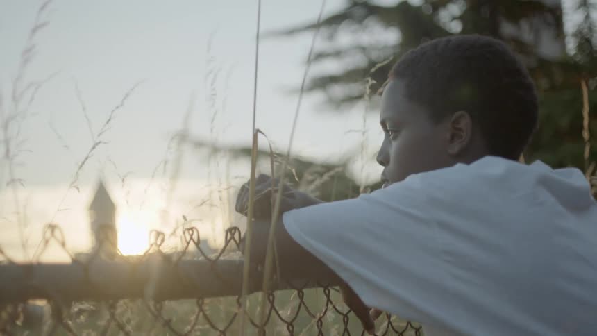A young boy in a white tee shirt leans against a chain link fence at sunset