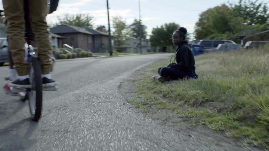 A girl sitting in the grass throwing rocks at a pair of kids riding a bicycle together in front of her