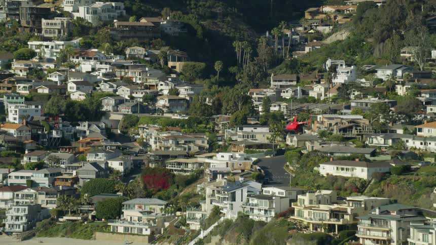 A red helicopter flies over a neighborhood of homes on a hillside with palm trees and trees