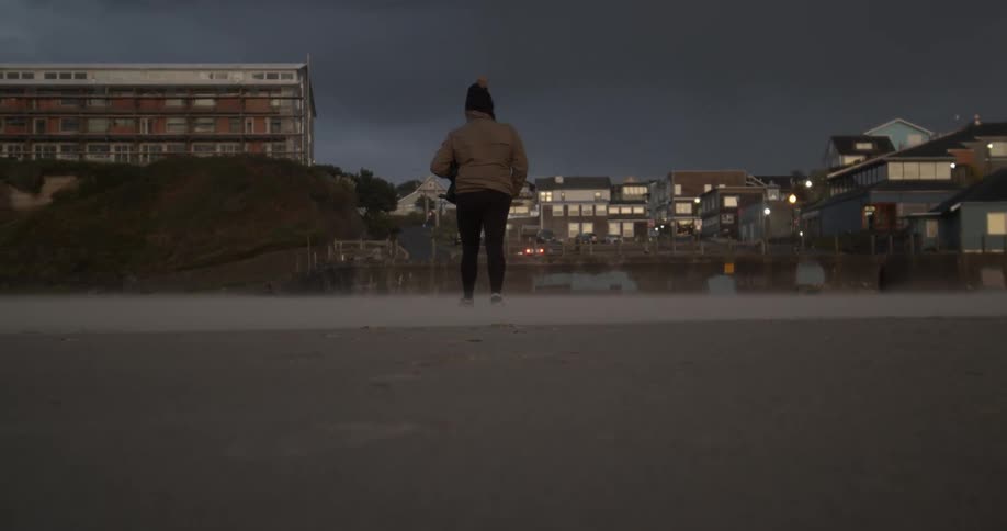 A woman wearing leggings and a coat walks on the sand towards the buildings and lights on shore