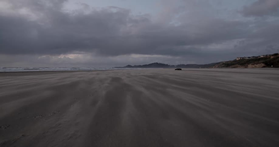 Car drives across wide sandy beach near town perched on low mountain