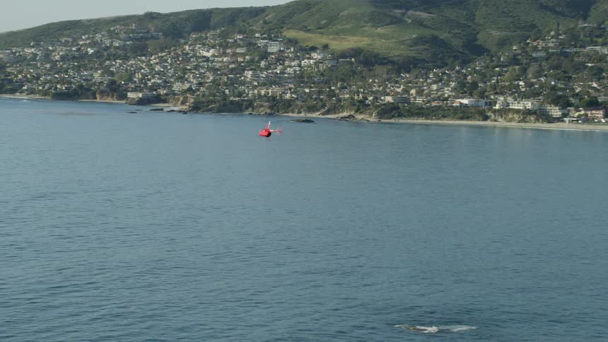 A red helicopter flies over the ocean off the coast of a city