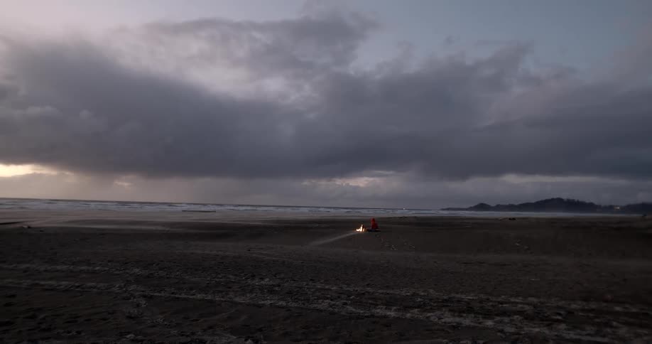 Lone person in distance near a bonfire at sunset under stormy clouds at the beach