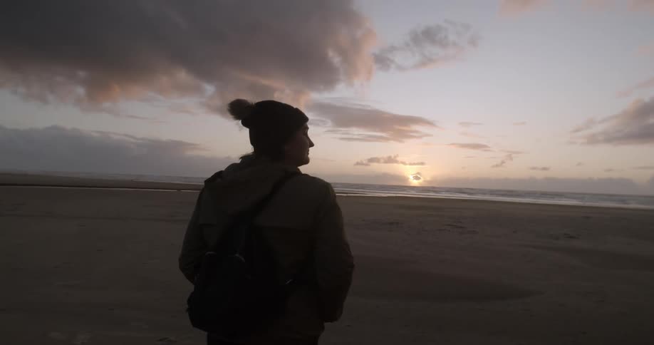 A person with a hat and a backpack walking on a sandy beach with the sky in the foreground