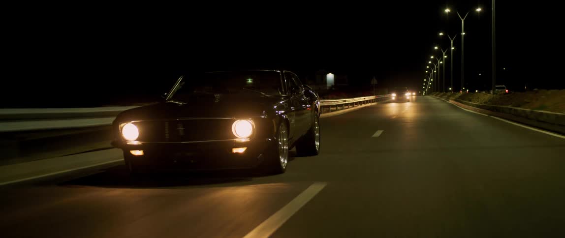 A black muscle car travels on a two lane road with headlights glowing at night