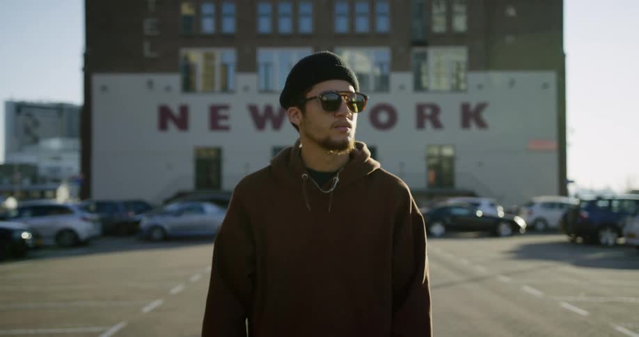 Man wearing hoodie and sunglasses walks through parking lot at sunrise