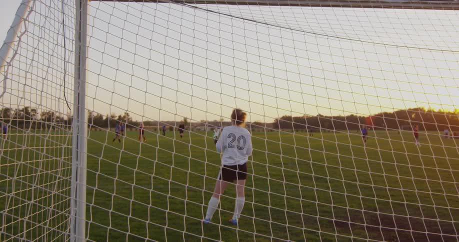 A goalie is holding a soccer ball by the goal as the sun sets
