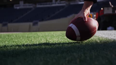 A hand reaches down and picks up a football off a field in a stadium