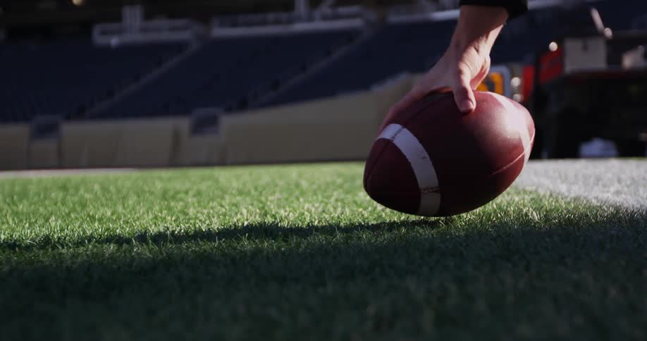 A hand reaches down and picks up a football off a field in a stadium