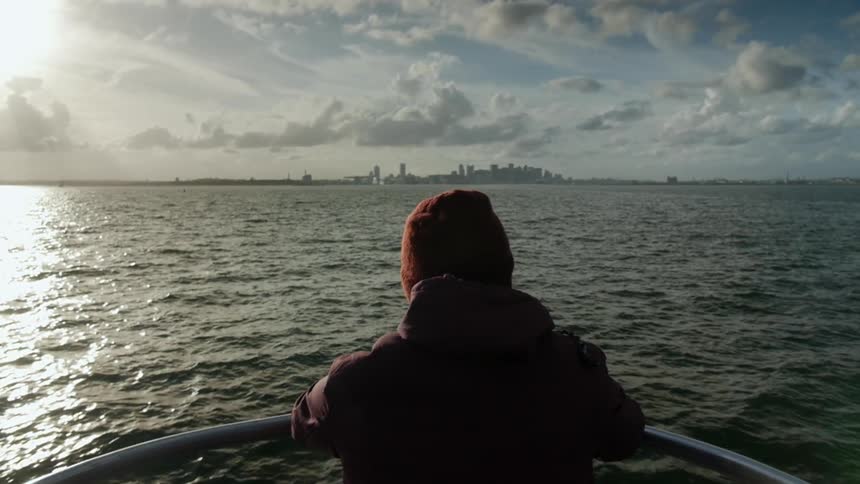 Person stands at boat railing as a city skyline approaches