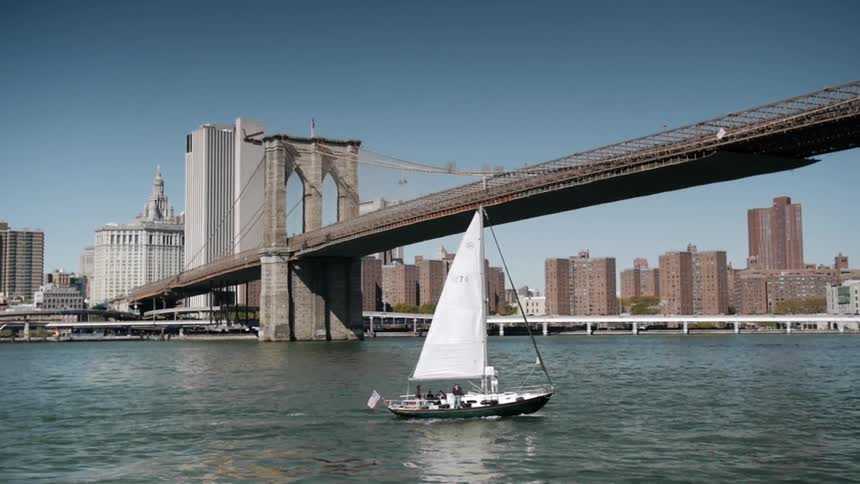 A boat with white sails floats under a bridge