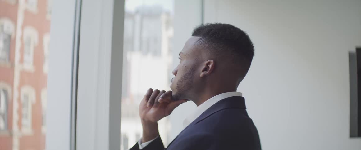 A black man standing in front of a window in an office while lost in thought