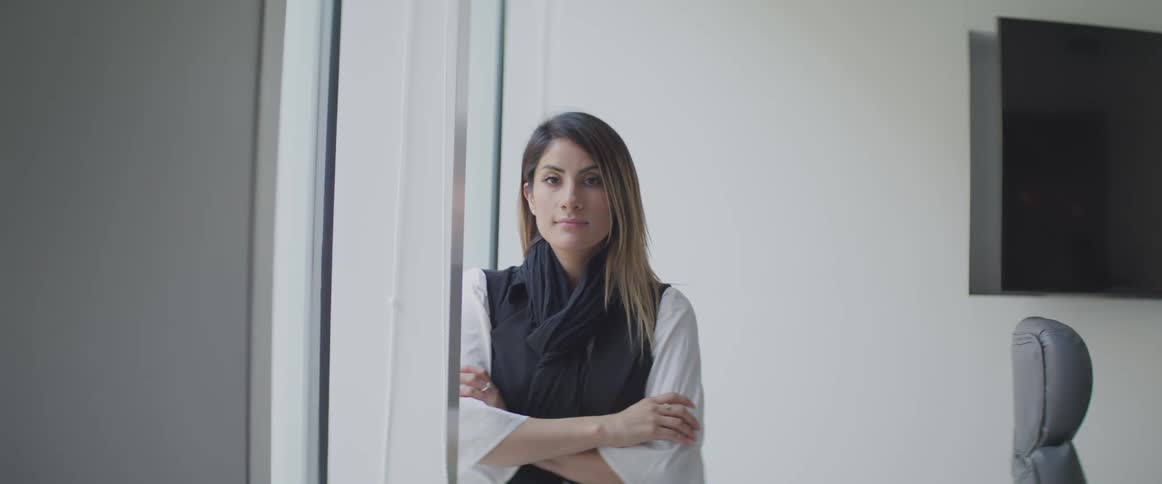 A woman stands with her arms crossed next to a large window in an office conference room