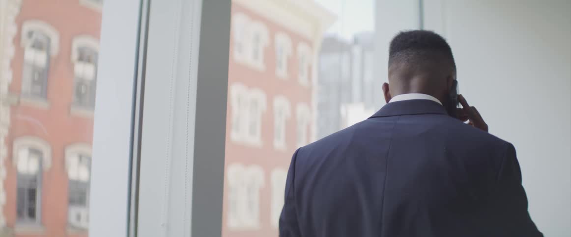 Man wearing business suit walks next to large windows talking on mobile phone