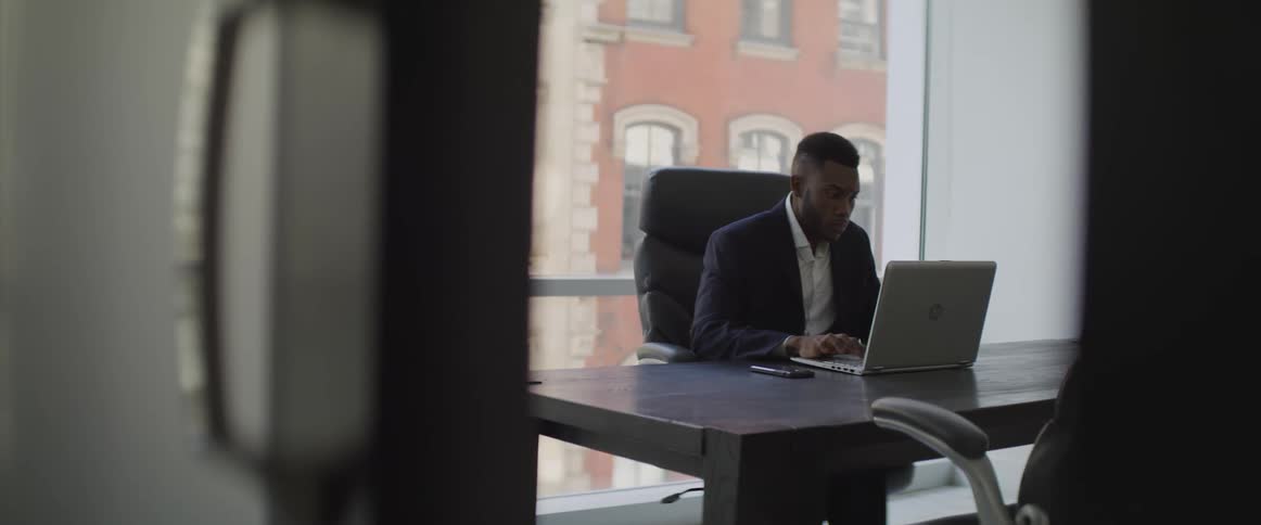 A black man works on a laptop at a desk in an office