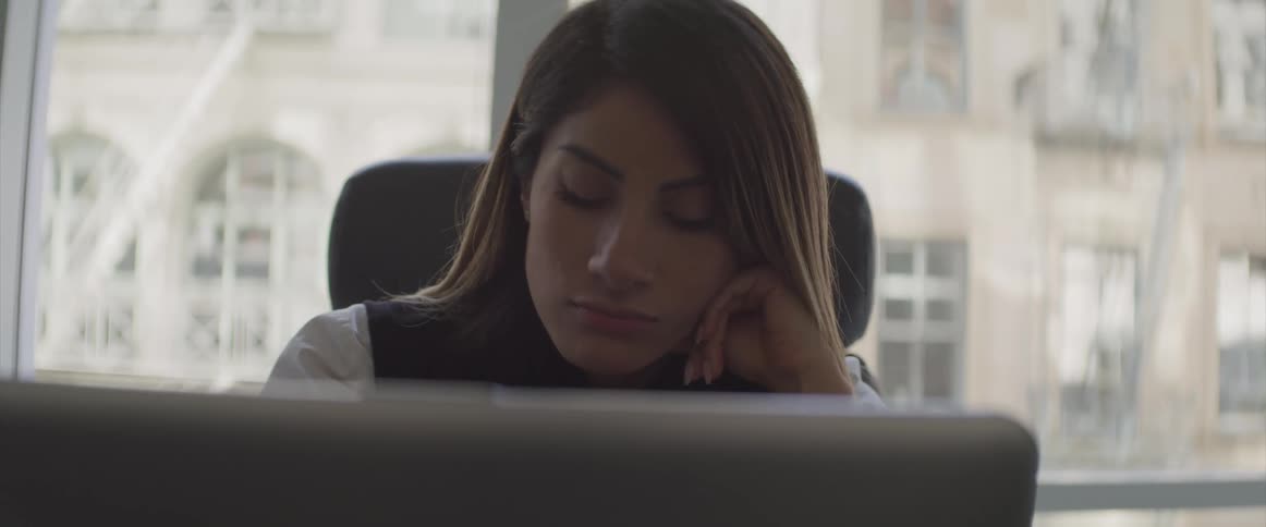 A woman sits in her office at her computer and is falling asleep