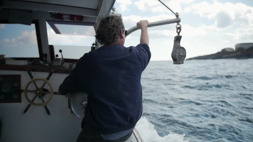 A man steers a fishing boat while sitting on the side