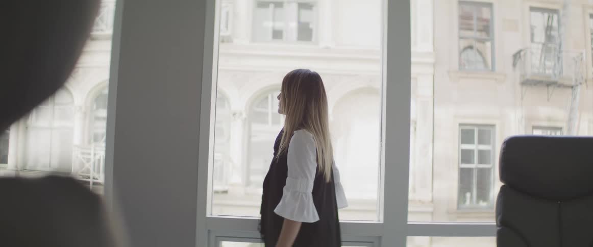 Female stands at the window in her office and looks out into the neighborhood while remaining very still