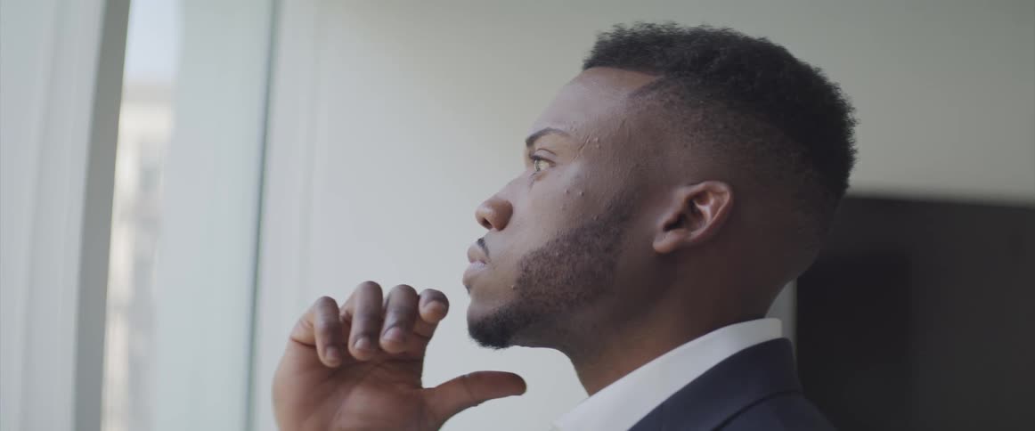 A man wearing business attire stands in an office in profile and lifts his hand to his chin as he thinks