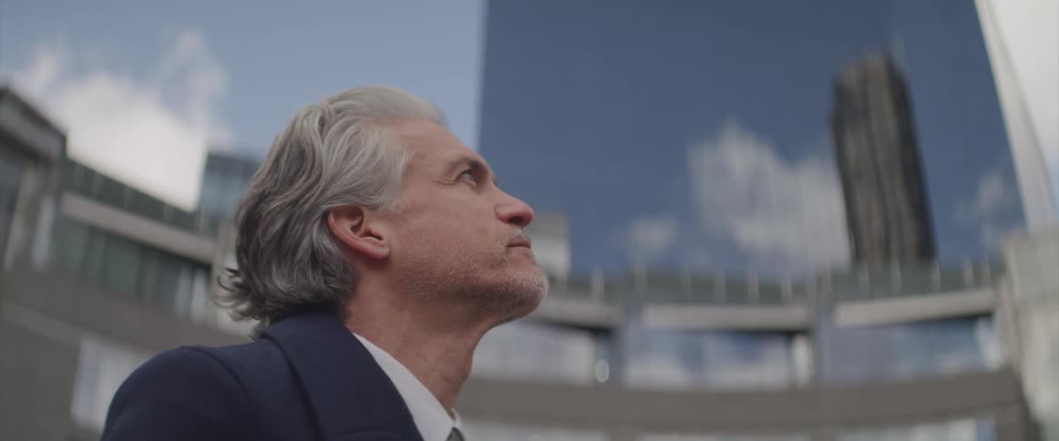 A graying man in a suit standing outside a large building and gazing at the sky