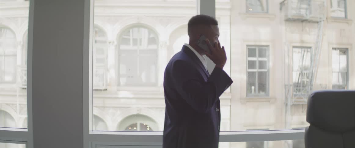 A man talks on the phone while walking past large windows in his executive office