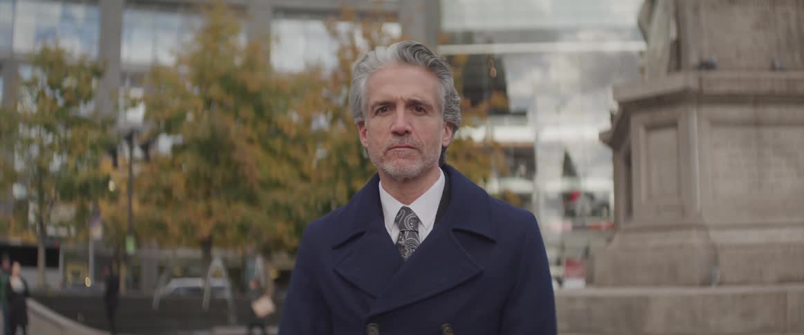 Man with gray hair and wearing a suit stands in the park and turns his head to face the camera