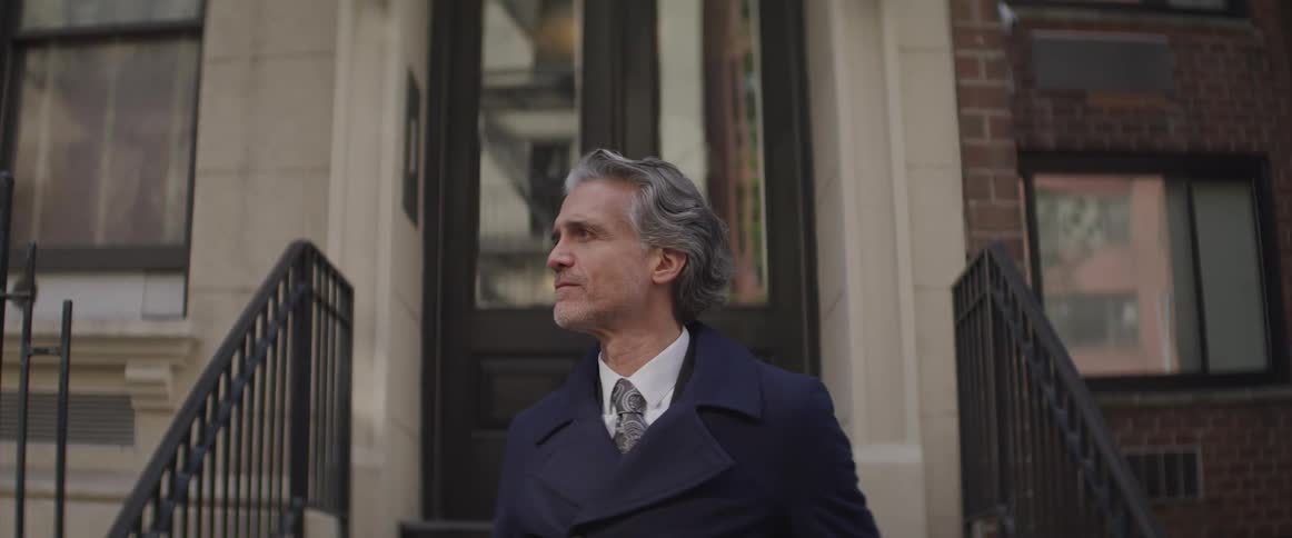 Man stands in front of stairway leading to a brownstone home looking down the street as he is waiting
