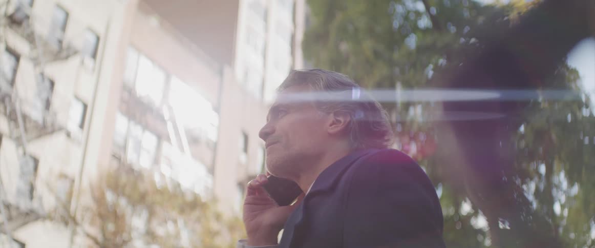 An older white man stands outdoor in a city with a phone up to his ear