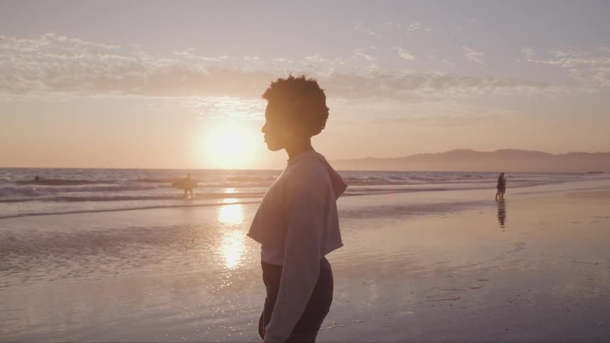 A woman stands where the ocean meets the beach as a surfer and a family frolic behind her