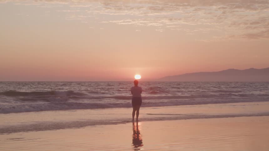 Woman standing on the wet sand beach watches the sunset next to the sea