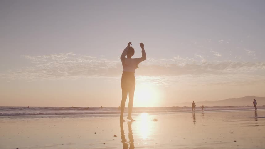 Black woman dances on the wet sand beach at sunset next to the sea