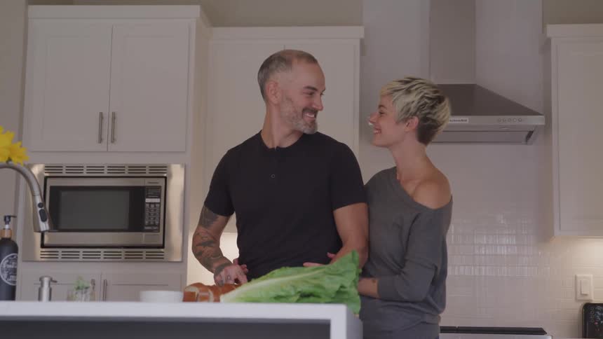 A man chops food in a home kitchen and his wife walks in beside him
