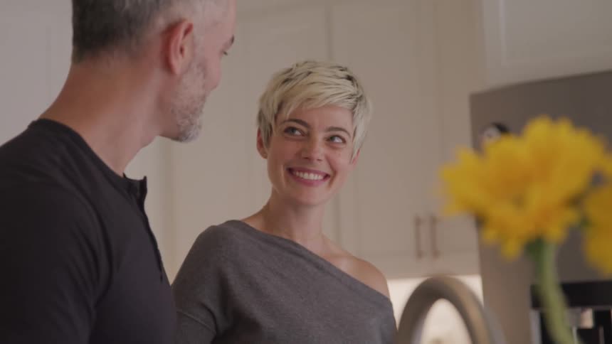 A man and a woman standing in a kitchen looking at each other and smiling