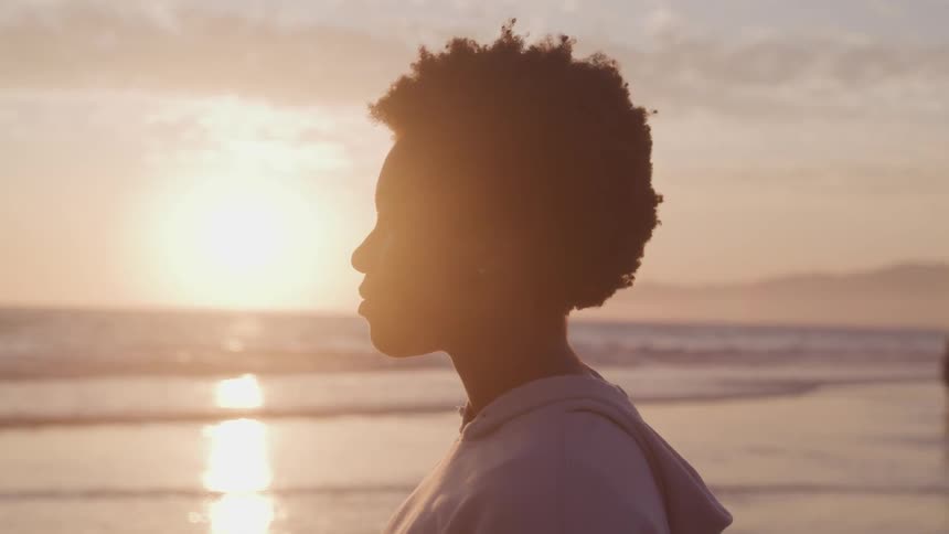 Black woman standing on the wet sand beach watches the sea at sunset