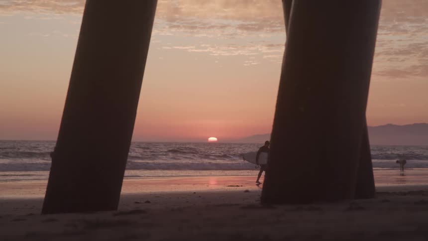 View from under pier of surfer walking in from the ocean at sunset