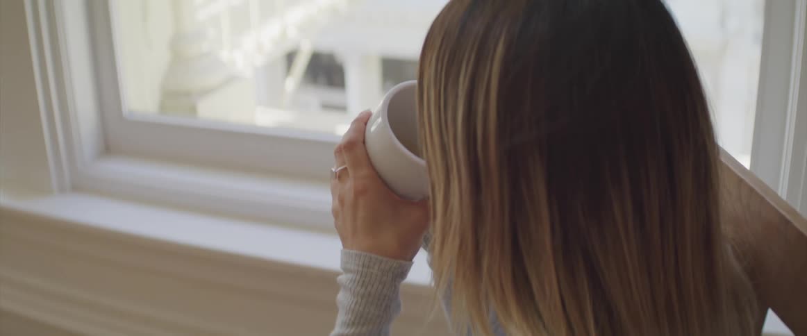 A woman sips a coffee mug held with both hands while sitting next to a large window