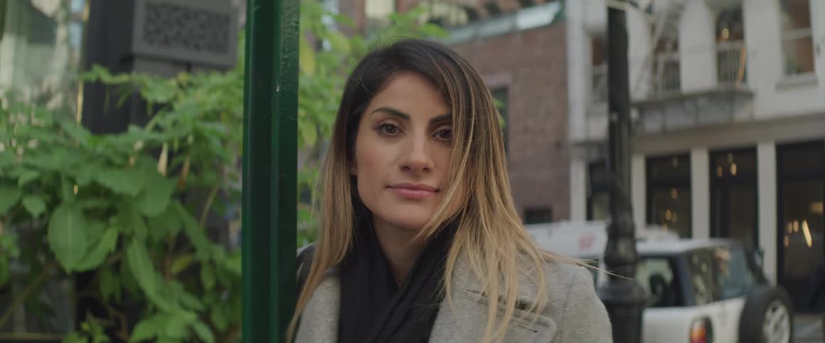 Female leaning on traffic sign pole is looking down and then raises her eyes to stare straight ahead without changing her neutral emotion